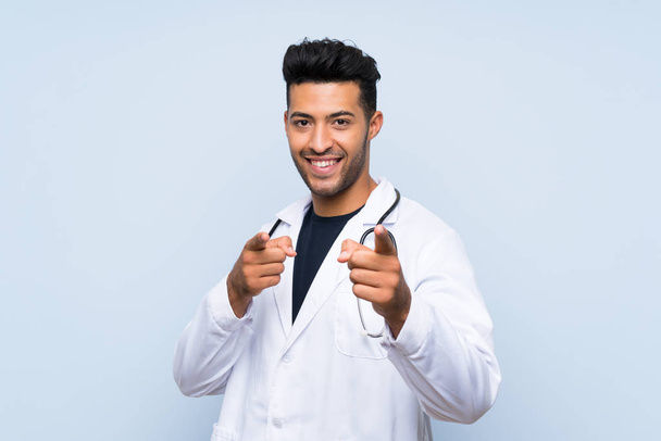Un joven doctor sobre una pared azul aislada te señala con el dedo con una expresión confiada
 - Foto, Imagen