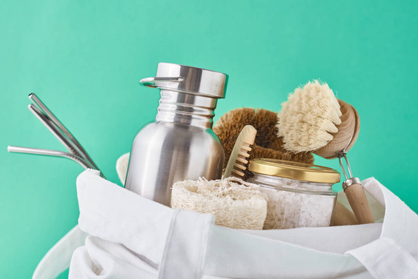 Reusable plastic free items in cotton shopping bag. Glass jar, metal straws, aluminum bottle and wooden cleaning brush on a green background - Photo, Image