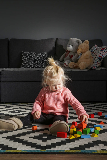 Adorable toddler girl is sitting on the floor and playing with colorful wooden blocks, natural light candid portrait - 写真・画像