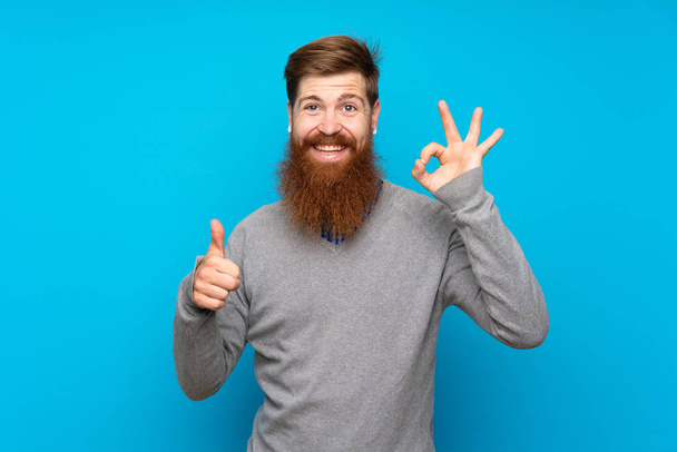 rothaariger Mann mit langem Bart vor isoliertem blauen Hintergrund, der Ok-Zeichen und Daumen-hoch-Geste zeigt - Foto, Bild