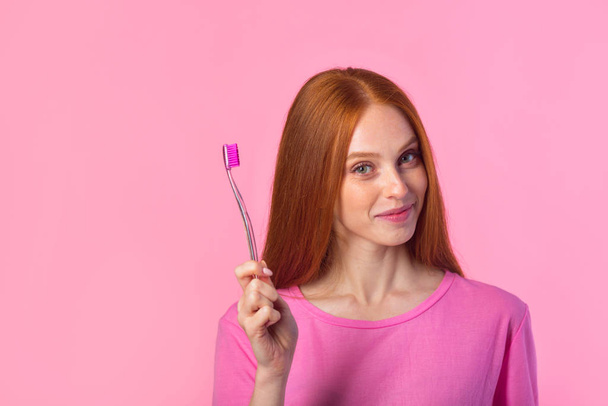 beautiful young woman with red hair on a pink background with a toothbrush - Foto, Imagen