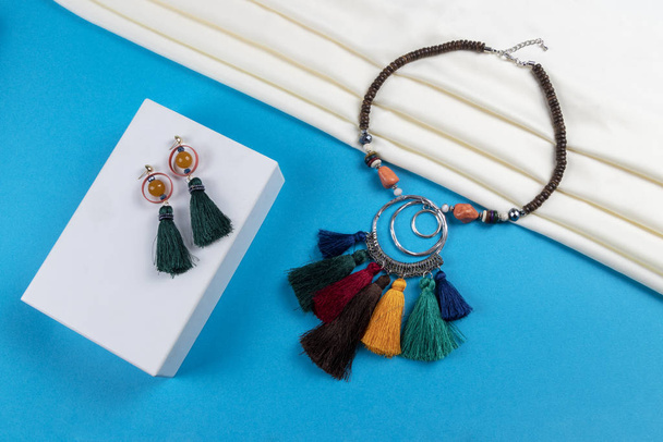 Set Quaste Ohrringe und Holzperlen mit bunten Quasten und Kristallperlen Halskette Boho-Stil auf weißem Schal und blauem Papierhintergrund - Foto, Bild