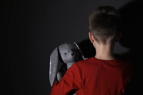 Sad little boy with toy near black wall, back view. Domestic violence concept - 写真・画像