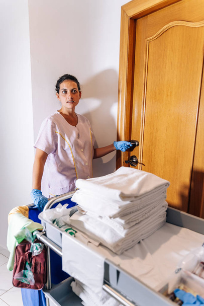 Foto vertical de un limpiador de hotel entrando en una habitación con un carro de limpieza y toallas limpias - Foto, Imagen
