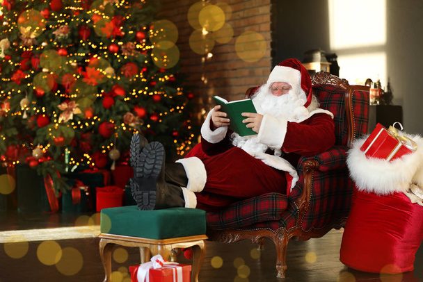 Santa Claus reading book near decorated Christmas tree indoors - Foto, afbeelding
