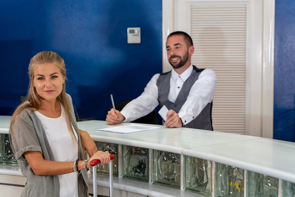 Stock photo of a young blonde girl at the entrance of a hotel with a suitcase and the receptionist standing waiting to attend her - Photo, Image