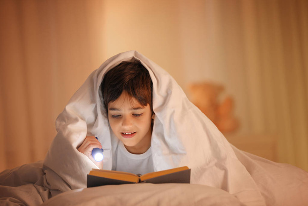 Boy with flashlight reading book under blanket at home - Фото, зображення
