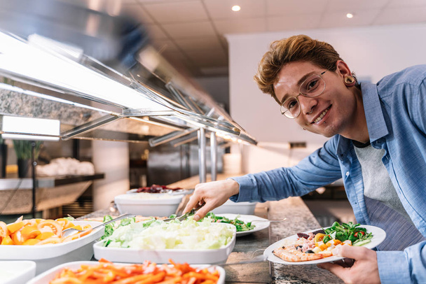 Stock photo of a young man with glasses and piercings taking food from a self-service - Photo, Image