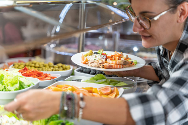 Foto de uma menina com óculos pegando comida com um prato de salada na mão em um balcão de restaurante self-service. Estilo de vida - Foto, Imagem