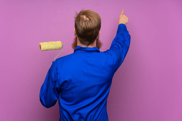 Painter man with long beard over isolated purple background pointing back with the index finger - Foto, afbeelding