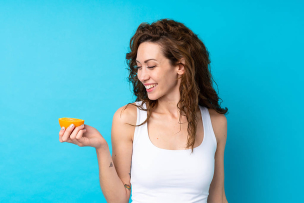 Young woman with curly hair holding an orange over isolated blue background - Foto, afbeelding