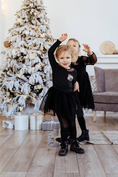 Two cute adorable little sisters near christmas tree in cozy living room. - Foto, afbeelding