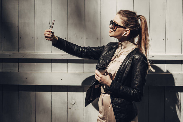 Portret stylowej dziewczyny nosi kobiet kombinezon i czarna skórzana kurtka pić kawę, korzystanie Smartphone - Zdjęcie, obraz