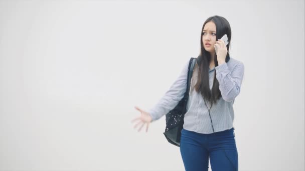Emotional young asian girl is on the phone, discussing some problems, waving her hands, looking upset. Backpack over her shoulder. - Кадри, відео
