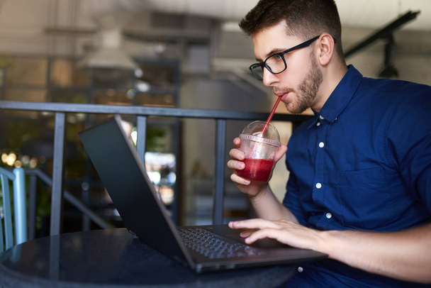Freelancer man with plastic cup of fresh beverage in hand works with laptop. Businessman in glasses drinks juice for body hydration while working. Attractive designer quench thirst. Healthy lifestyle. - Photo, Image