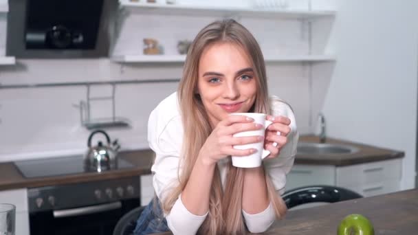 Cheerful blonde woman drinking morning coffee at the kitchen. Looking at the camera and smiling. Handheld shot. Slowmotion. - Video