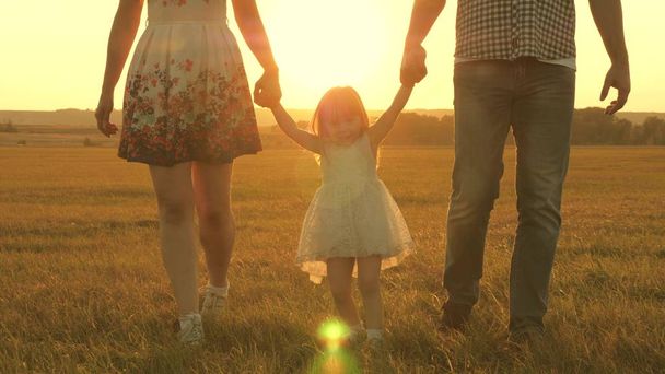 太陽の背景に公園でお父さんとお母さんの手を握ってジャンプ小さな娘。家族の概念。子供は夕日の光の中でフィールドでお父さんとお母さんと遊びます。自然の中で小さな子供と歩く. - 写真・画像