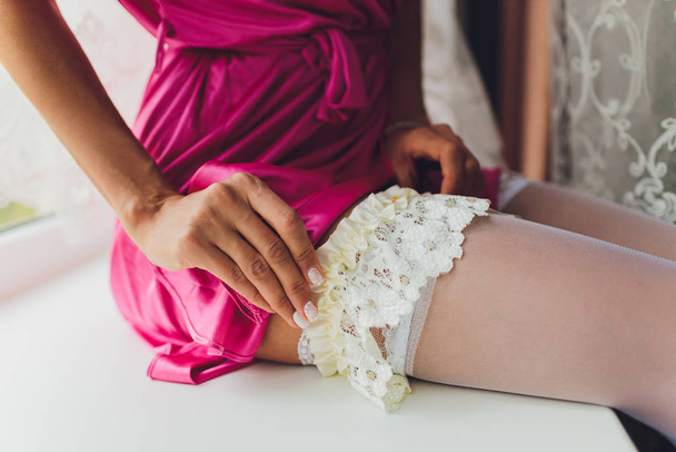 In the morning, the bride in stockings and a white wedding dress wears a garter on her leg, the bride is holding her hands for the garter. - 写真・画像