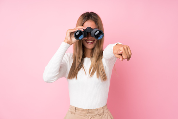 Young blonde woman over isolated pink background with black binoculars - Photo, Image
