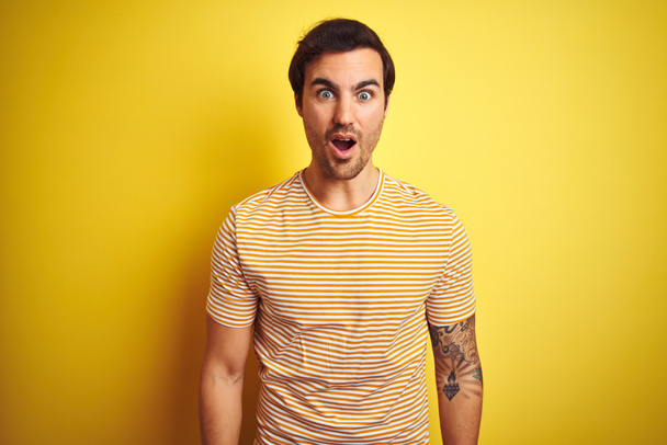 Młody przystojny mężczyzna z tatuażem w pasiastej koszulce na odizolowanym żółtym tle przerażony i zszokowany zaskakującą ekspresją, strachem i podnieconą twarzą. - Zdjęcie, obraz