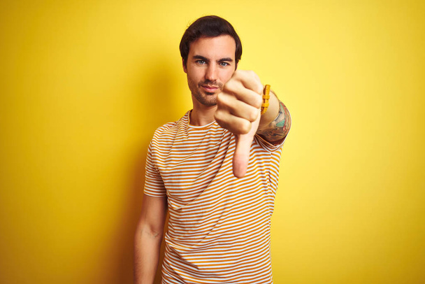 Young handsome man with tattoo wearing striped t-shirt over isolated yellow background looking unhappy and angry showing rejection and negative with thumbs down gesture. Bad expression. - Foto, Bild