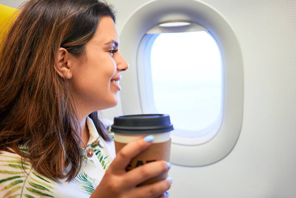 Jonge reiziger vrouw zitten in het vliegtuig op de luchthaven met uitzicht op de hemel uit het raam het drinken van een kopje koffie - Foto, afbeelding