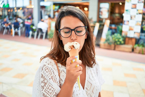 Nuori kaunis nainen syö jäätelötötteröä ostoskeskuksessa aurinkoisena kesäpäivänä lomilla.
 - Valokuva, kuva