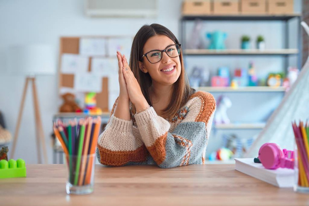 Młoda piękna nauczycielka ubrana w sweter i okulary siedząca na biurku w przedszkolu klaskając i bijąc brawo szczęśliwym i radosnym, uśmiechniętym dumnym dłoniom razem - Zdjęcie, obraz