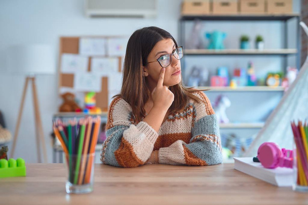 Young beautiful teacher woman wearing sweater and glasses sitting on desk at kindergarten Pointing to the eye watching you gesture, suspicious expression - Photo, Image