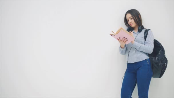 Beautiful asian young teenager high school student girl standing with backpack behind shoulders headphones on neck, reading a book. - Séquence, vidéo