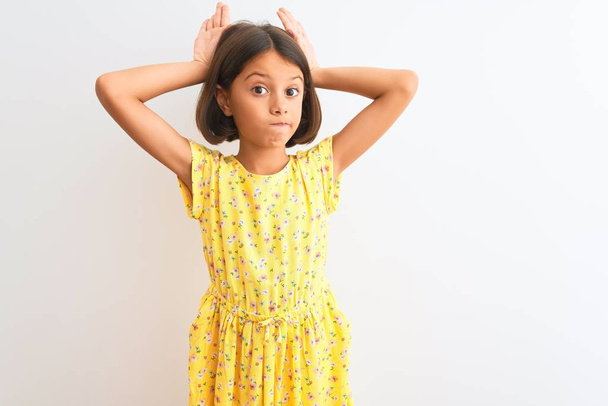 Young beautiful child girl wearing yellow floral dress standing over isolated white background Doing bunny ears gesture with hands palms looking cynical and skeptical. Easter rabbit concept. - Photo, Image