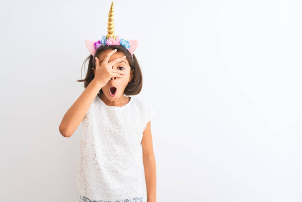 Beautiful child girl wearing unicorn diadem standing over isolated white background peeking in shock covering face and eyes with hand, looking through fingers with embarrassed expression. - Foto, Bild