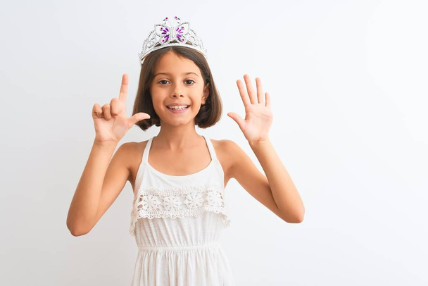 Gyönyörű gyermek lány visel hercegnő korona áll az elszigetelt fehér háttér mutatja, és rámutatva ujjak száma 7 miközben mosolygós magabiztos és boldog. - Fotó, kép