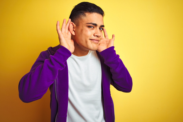 Young brazilian man wearing purple sweatshirt standing over isolated yellow background Trying to hear both hands on ear gesture, curious for gossip. Hearing problem, deaf - Foto, imagen