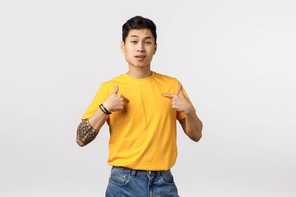 ehrgeizige gut aussehende asiatische männliche Snob in gelbem T-Shirt, mit Tätowierungen, zeigt sich mit selbstbewusstem, machohaftem Gesichtsausdruck, lobt seine eigenen Fähigkeiten, prahlt, wie cool und genial er ist, weißer Hintergrund - Foto, Bild