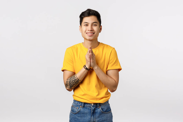 Милий дружній і приємний молодий азіатський татуйований хлопець з милою посмішкою, тримає руки разом біля грудей в жесті молитви, благаючи про послугу, благаючи або подякуючи, білий фон
 - Фото, зображення