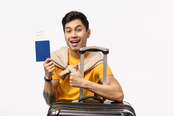 Neşeli, kaygısız yakışıklı Asyalı adam, siyah bavulun yanında oturuyor, onaylıyor, pasaport ve uçak bileti tutuyor, mutlu bir şekilde gülümsüyor, yurtdışında mükemmel bir tatil için seyahat acentesinde turne satın alıyor. - Fotoğraf, Görsel