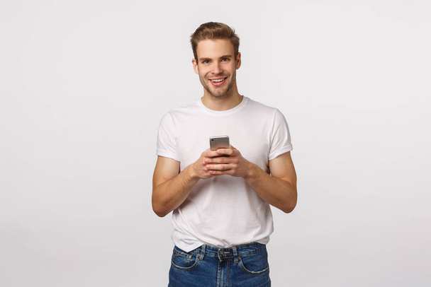 Telefon, digitales modernes Leben, Technologiekonzept. schöner lächelnder blonder kaukasischer Mann mit Borsten, trägt weißes T-Shirt und Jeans, hält das Smartphone in der Hand, als plaudere er mit der Kamera - Foto, Bild