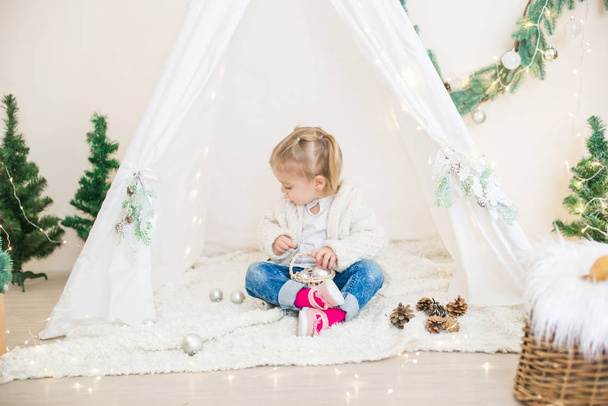 Little cute girl in a white warm knitted cardigan near a children's lodge decorated with a garland and needles, toys and tangerines. Christmas mood - Foto, afbeelding