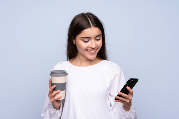 Jeune femme sur fond bleu isolé tenant café à emporter et un mobile
 - Photo, image