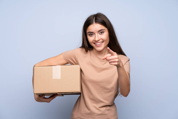 Young woman over isolated blue background holding a box to move it to another site while pointing to the front - Photo, Image