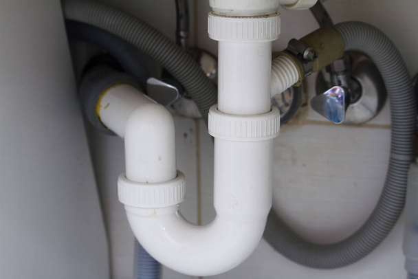 Ловушка PVC серый под кухней раковина из нержавеющей стали с гибкой трубки над потоком подключения в белой стене керамической плитки
 - Фото, изображение