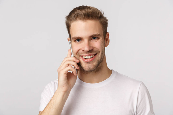 Γεια σου Ιμ. Φιλικός όμορφος χαμογελαστός ξανθός καυκάσιος με λευκό μπλουζάκι απαντώντας, κρατώντας το smartphone κοντά στο αυτί, μιλώντας, κάνοντας τάξη, στέκεται λευκό φόντο - Φωτογραφία, εικόνα
