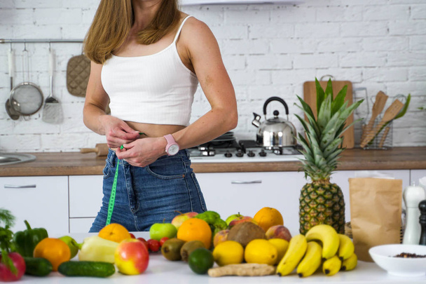 A vegan girl in the kitchen near the fruit measures her waist.  - Foto, afbeelding