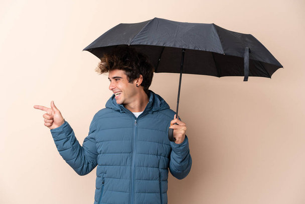 Hombre sosteniendo un paraguas sobre un fondo aislado apuntando hacia un lado para presentar un producto
 - Foto, Imagen