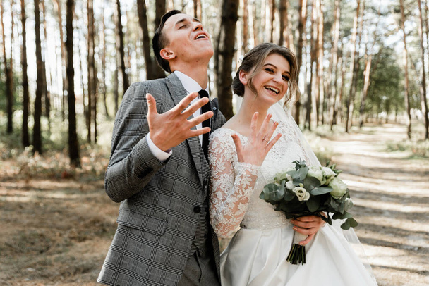 Attractive couple celebrating their wedding in forest. Portrait of young happy groom and bride in wedding clothes standing together, holding hands and looking at each other. Wedding ring - Photo, Image