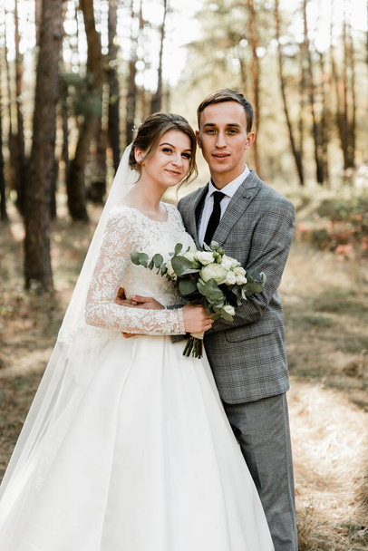 Attractive couple celebrating their wedding in forest. Portrait of young happy groom and bride in wedding clothes standing together, holding hands and looking at each other - Photo, Image