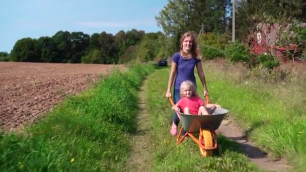 Liefdevolle vrouw duwen kruiwagen met gelukkig klein meisje door dorp manier - Video