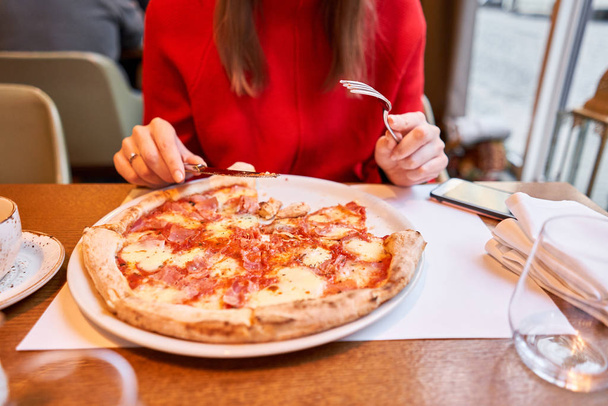 Kobieta je nożem i pizzę capricciosa. Pizza z pomidorami mozzarelli, karczochem, czarnymi oliwkami i bazylią. Neapolitańska pizza z pieca opalanego drewnem. obiad we włoskiej restauracji. - Zdjęcie, obraz