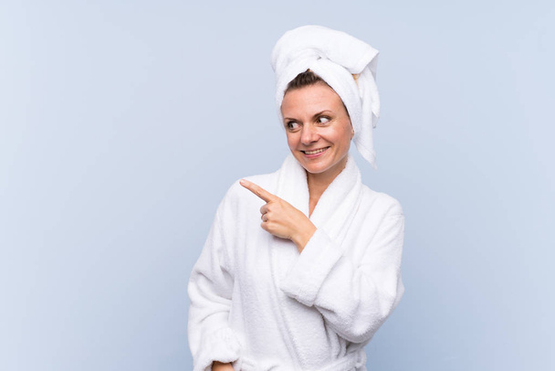 Woman in bathrobe over isolated blue background pointing finger to the side - Photo, Image